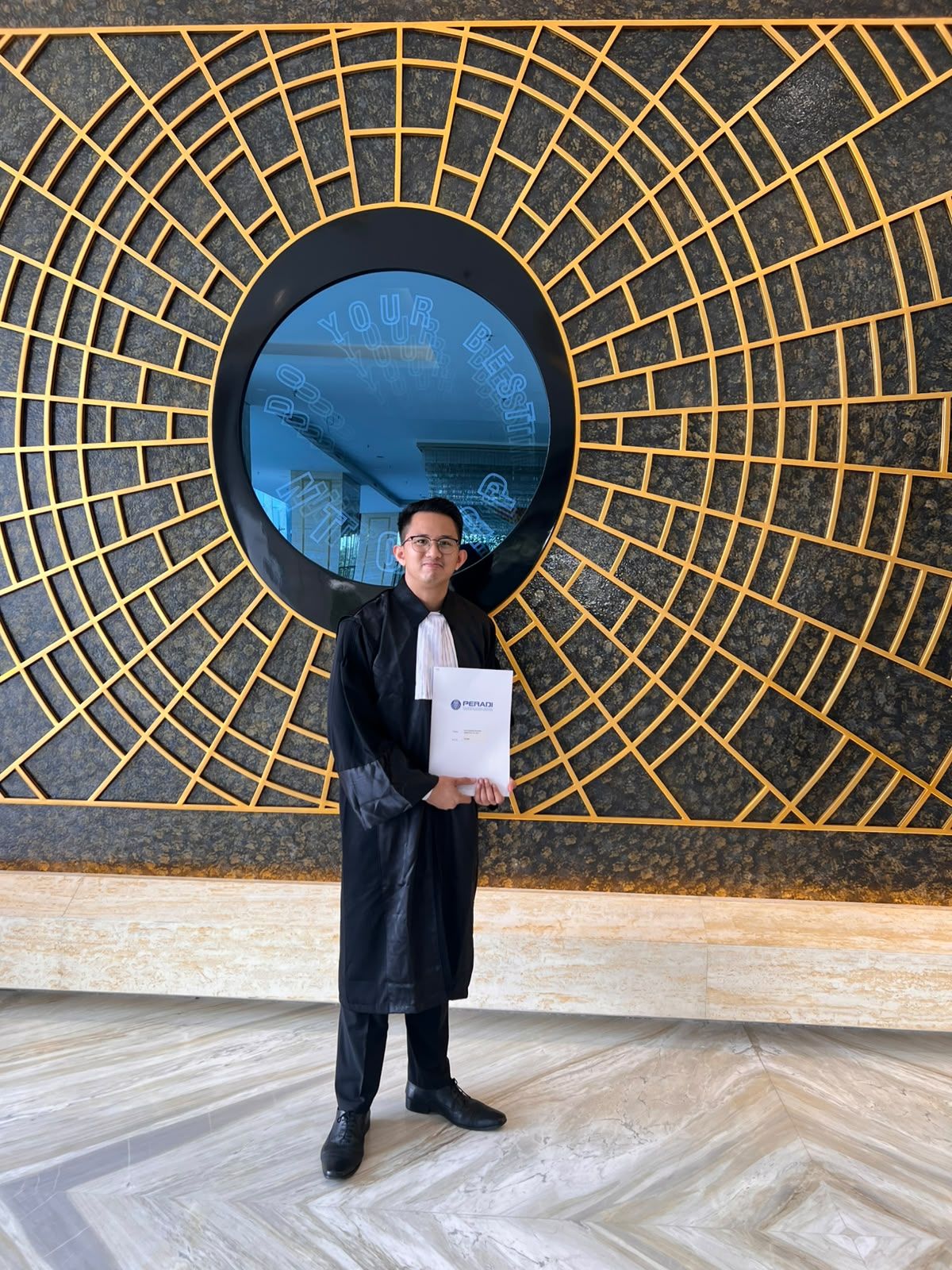 Dr. (Cand.) Putu George Matthew Simbolon, S.H., M.H. in advocate robes before a gold sunburst wall, holding his PERADI papers