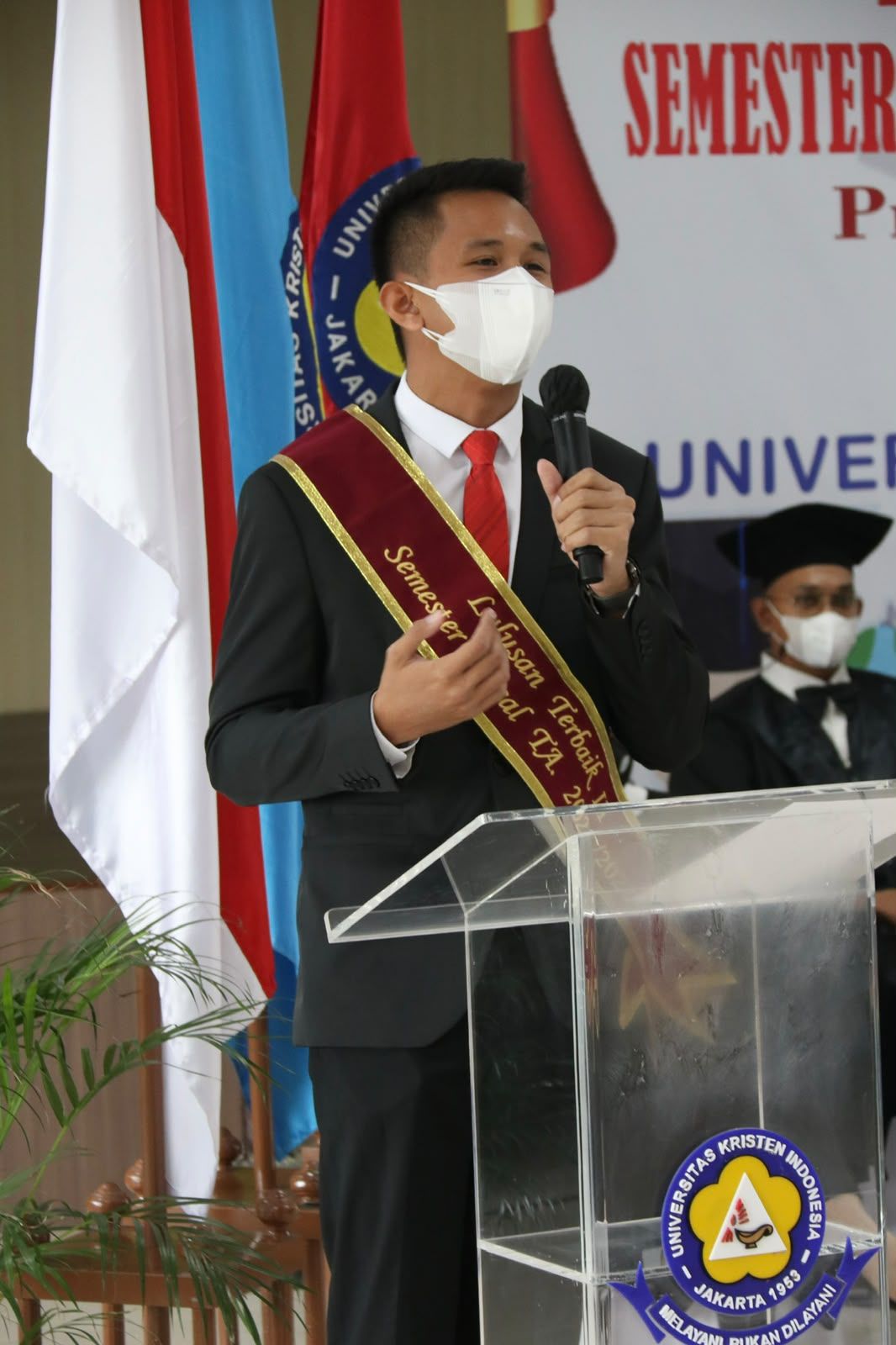 Dr. (Cand.) Putu George Matthew Simbolon, S.H., M.H. speaking at the Universitas Kristen Indonesia graduation, wearing a Summa Cum Laude sash