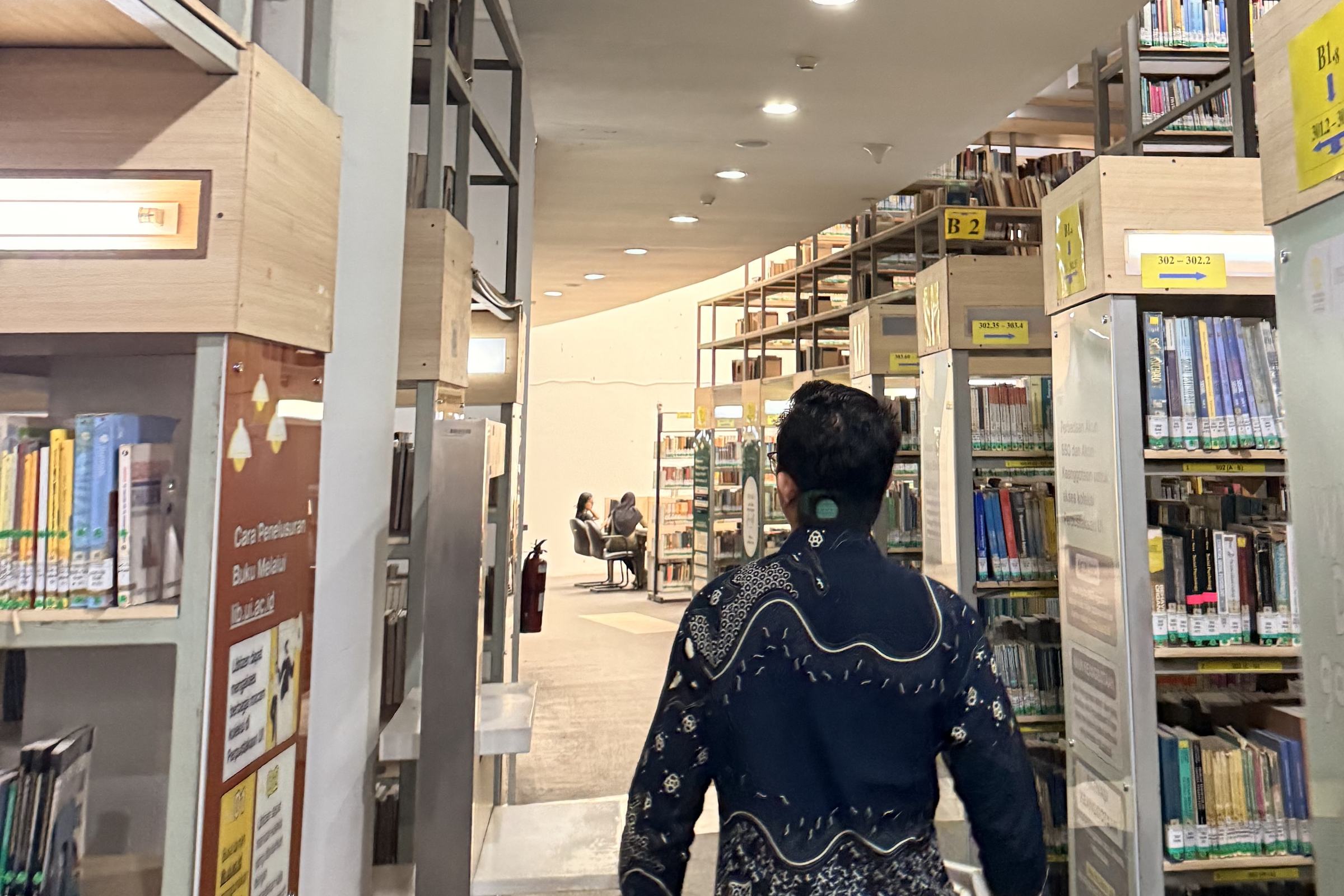 Dr. (Cand.) Putu George Matthew Simbolon, S.H., M.H. walking through the stacks of a law library, past a sign reading When in doubt, go to the library